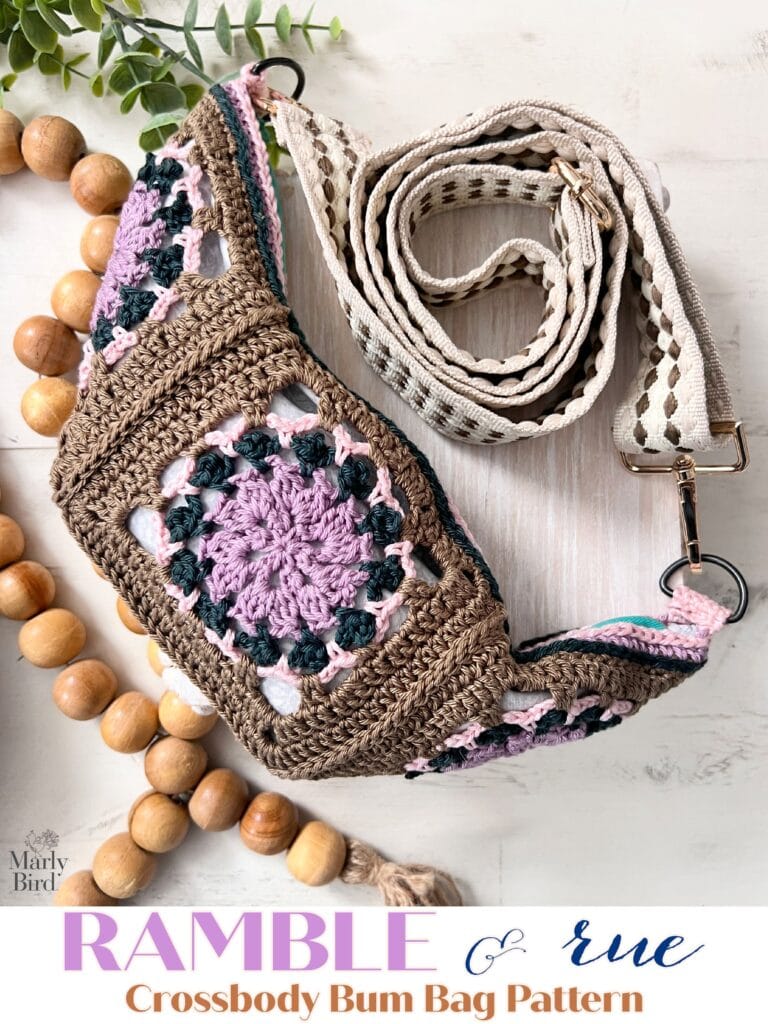 A crocheted crossbody bum bag with a granny square design in brown, purple, pink, and green yarn, displayed with its woven strap and wooden bead accent on a light surface. Text: "Ramble & Rue Crossbody Bum Bag Pattern. -Marly Bird