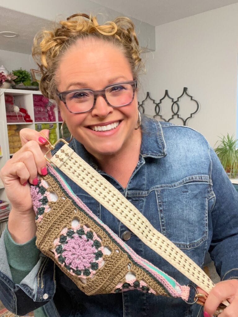 A smiling woman with curly hair and glasses holds up a colorful crochet crossbody bag pattern. She is wearing a denim jacket and standing in a bright, craft-filled room. -Marly Bird