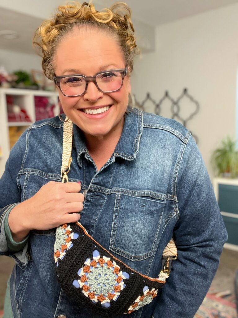 A smiling person with curly blonde hair, wearing glasses and a denim jacket, holds a crochet crossbody bag pattern featuring colorful granny squares in a cozy, well-lit room. -Marly Bird