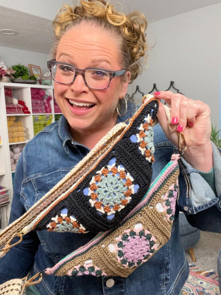 A smiling woman with glasses and curly hair holds up two colorful, crocheted bags in a room filled with yarn and crafting supplies, surrounded by inspiration from free knitting and crochet patterns. -Marly Bird