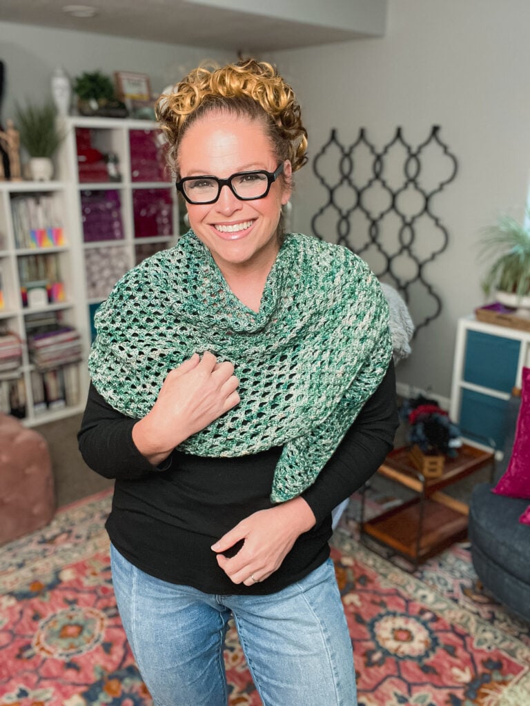 Marly Bird models a free knit boomerang shawl pattern. 
It is textured green crocheted shawl in a cozy room with books and plants, highlighting the shawl’s drape and stitch detail.