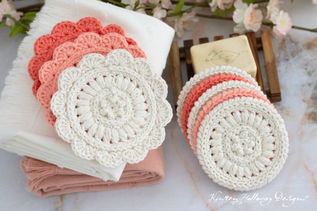 Crocheted coasters in pink and cream sit neatly stacked on a marble surface, echoing the essence of spring cleaning. Nearby, folded napkins in matching shades accompany a bar of soap on a wooden holder, while pink flowers add a touch of elegance to the background. -Marly Bird