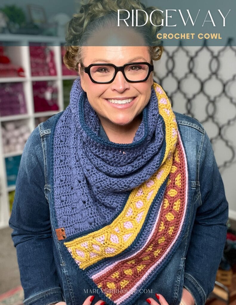 A smiling woman wearing glasses and a denim jacket models a colorful crochet cowl in blue, yellow, pink, and burgundy tones. The text reads "RIDGEWAY Crochet Cowl," perfect for the Marly Bird Turkey Trot Make-Along 2025. Yarn shelves are visible behind her. -Marly Bird