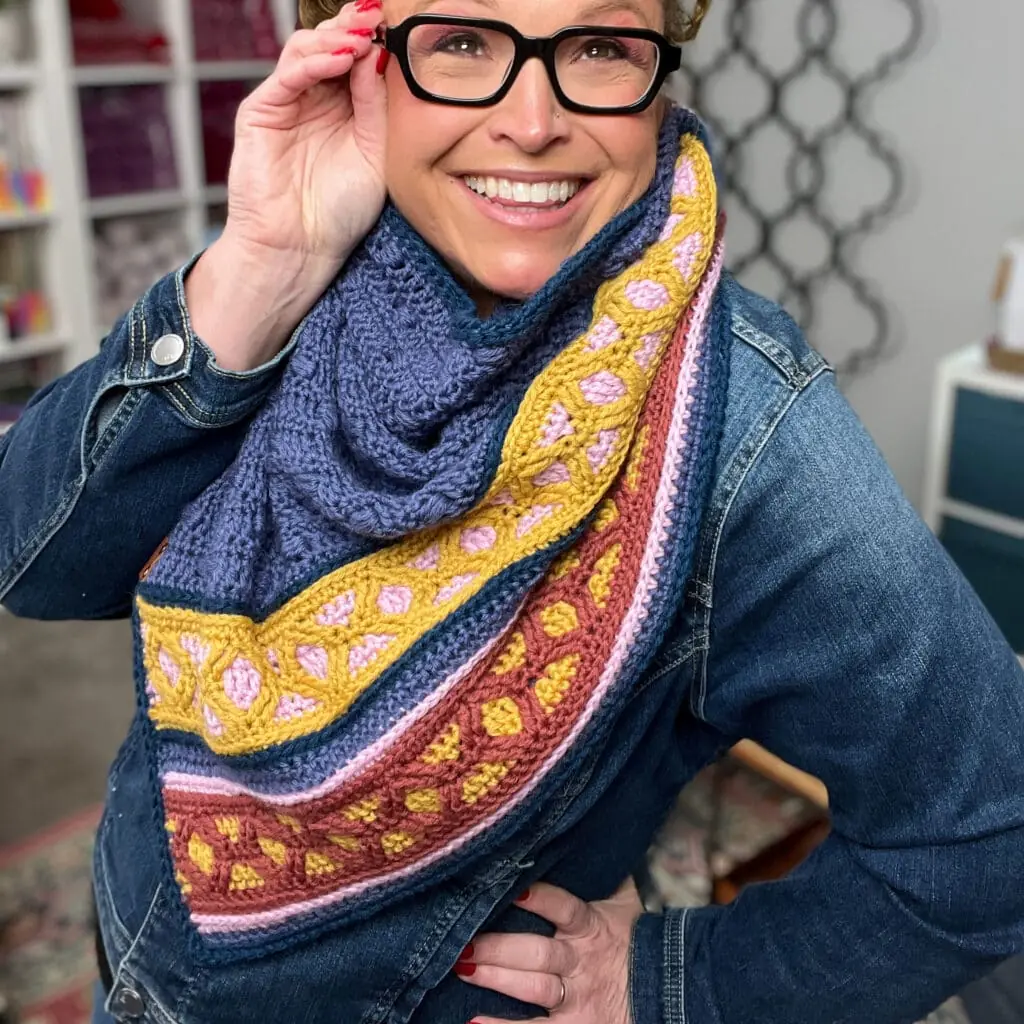 A smiling person wearing glasses and a blue denim jacket poses indoors, showcasing a colorful knitted scarf inspired by the Marly Bird Turkey Trot Make-Along 2025. The background features shelves filled with yarn. -Marly Bird