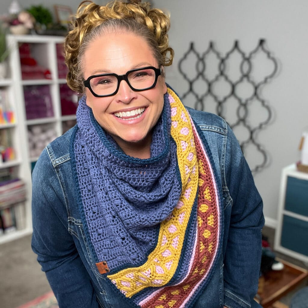 A smiling person with curly hair, glasses, and a denim jacket wears a colorful crocheted scarf, perhaps inspired by the Marly Bird Turkey Trot Make-Along 2025. Shelves with yarn and craft supplies are visible in the cozy room’s background. -Marly Bird