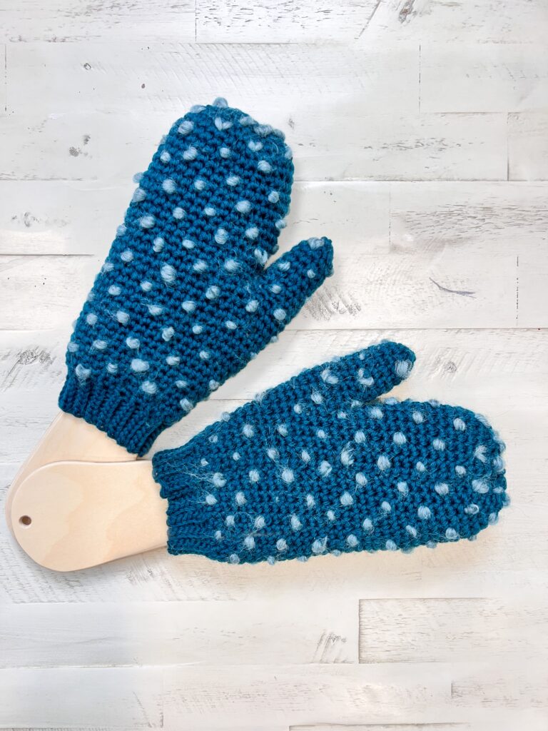 A pair of teal Andes Thrummed Knit Mittens with a bobble texture and white speckles are displayed on a white wooden surface, one mitten gently fitted on a wooden form. Find the Free Knitting Pattern in First Fall 2025. -Marly Bird