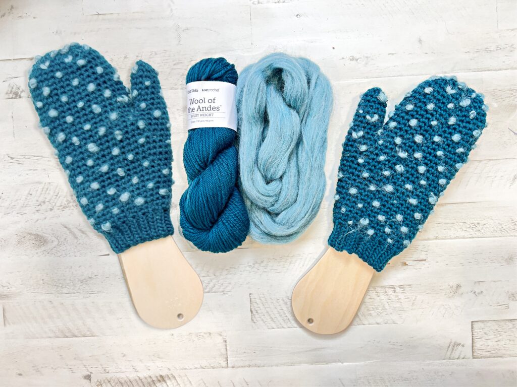 A pair of blue knit mittens with white dots on wooden blockers, a skein of blue yarn, and a bundle of light blue fiber evoke cozy Andes style—all arranged on a light wooden surface. Discover the free knitting pattern for these charming mittens. -Marly Bird