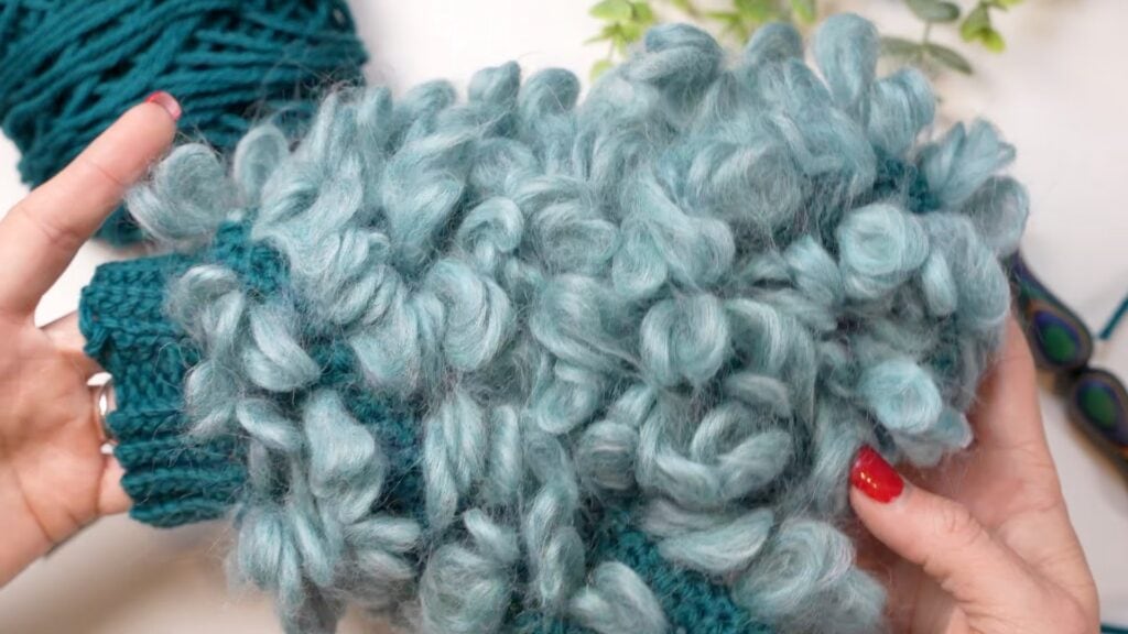 Hands with red nail polish hold a piece of chunky, teal yarn knitting in large, fluffy loops—perfect for a Fleece-Lined Crochet project. A ball of yarn, green plant, and scissors rest on the white surface behind. -Marly Bird