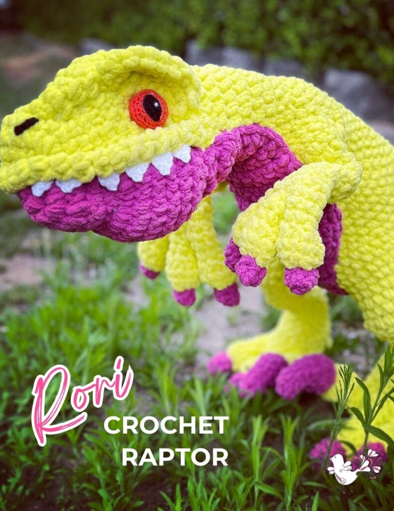 A vibrant crochet dinosaur plush with a bright yellow body and purple details is posed in a garden setting. The text reads "Rori the Raptor" in bold letters, showcasing this delightful crochet pattern. -Marly Bird