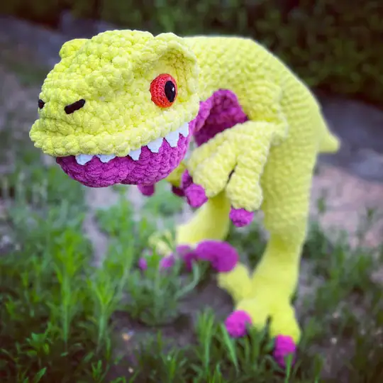 A plush, crocheted dinosaur toy with a bright yellow body, purple claws and mouth, and a red and black eye stands in green grass; perfect inspiration for those seeking free knitting and crochet patterns. -Marly Bird