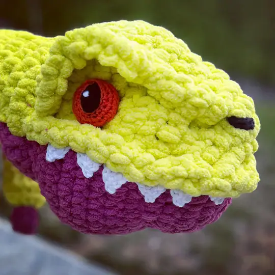 A close-up of a crocheted dinosaur head with bright yellow and purple yarn, red and black eyes, white felt teeth, and a playful expression showcases inspiration for free knitting and crochet patterns. The background is blurred greenery. -Marly Bird