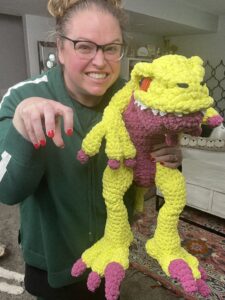 A person with glasses playfully poses with a charming crocheted yellow and pink dinosaur plush, mimicking its claws like a raptor. They stand in a room with a gray carpet and light-colored furniture, embodying the fun spirit of the crochet pattern. -Marly Bird