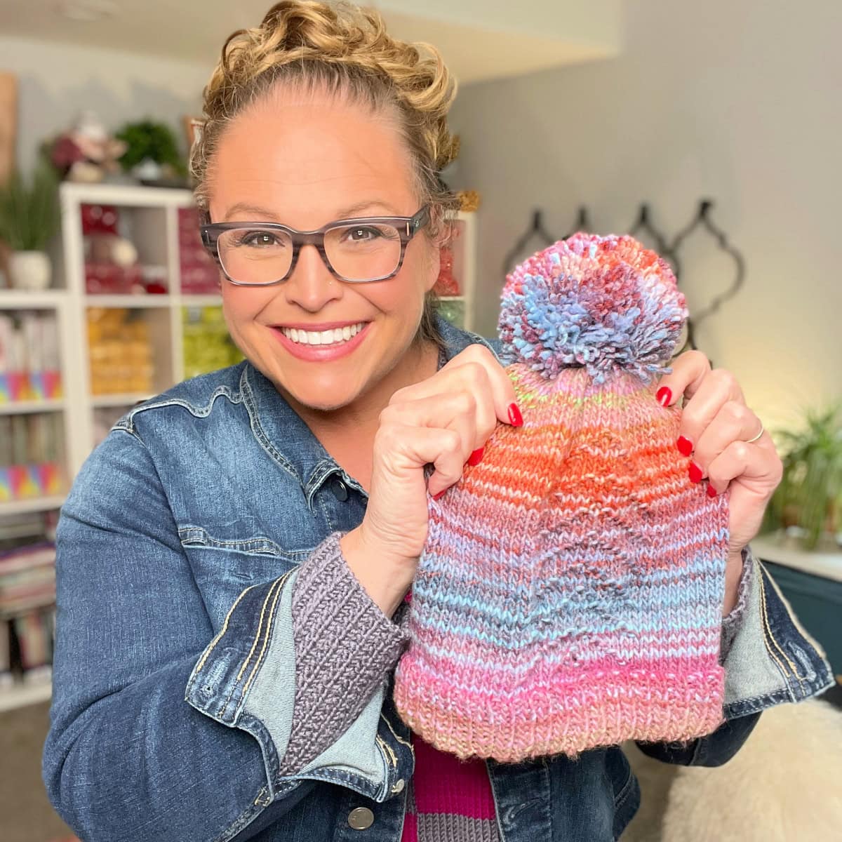 A smiling person wearing glasses and a denim jacket holds up a colorful knit hat pattern with a pom-pom, standing indoors in a bright, cozy room. -Marly Bird