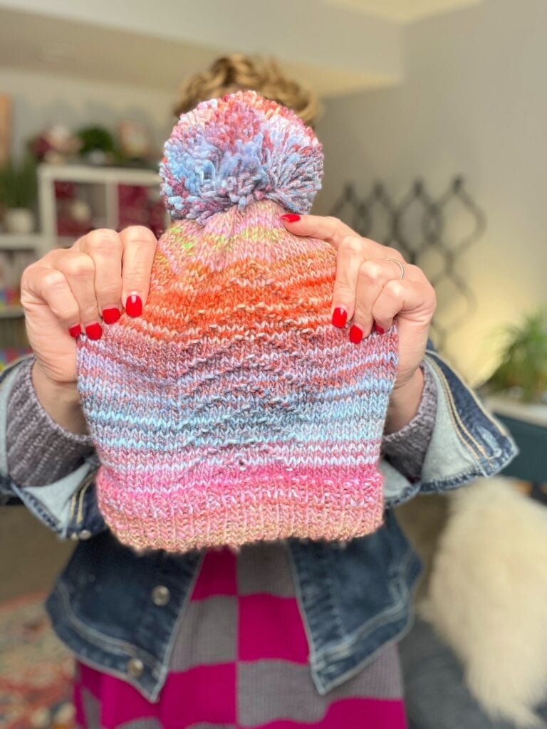 A person with red nail polish holds up a Roy G Biv Knit Hat, made from gorgeous yarn, while wearing a denim jacket over a pink and gray striped shirt. The cozy indoor background features plants and shelves. -Marly Bird