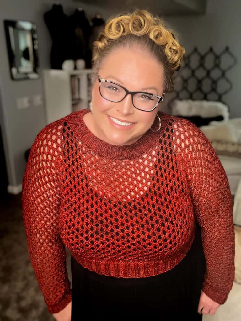 A person with glasses and styled blonde hair smiles warmly, wearing a red crochet-stitched top over a black undershirt. The background shows a dimly lit room with a sofa, a black decorative screen, and some shelves. -Marly Bird