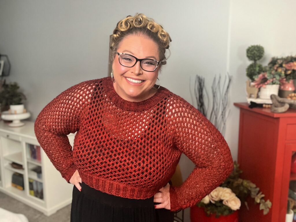 A person with blond curly hair, wearing glasses and a red crocheted sweater, is smiling at the camera. They are indoors with a red cabinet and various decor items, including flowers and books, in the background. -Marly Bird