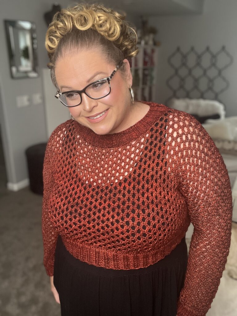 A person wearing glasses and a red crochet sweater over a black top stands indoors, smiling. They have blonde hair styled in curls. The background includes a gray wall with picture frames, a white sofa, and a decorative wall piece. -Marly Bird