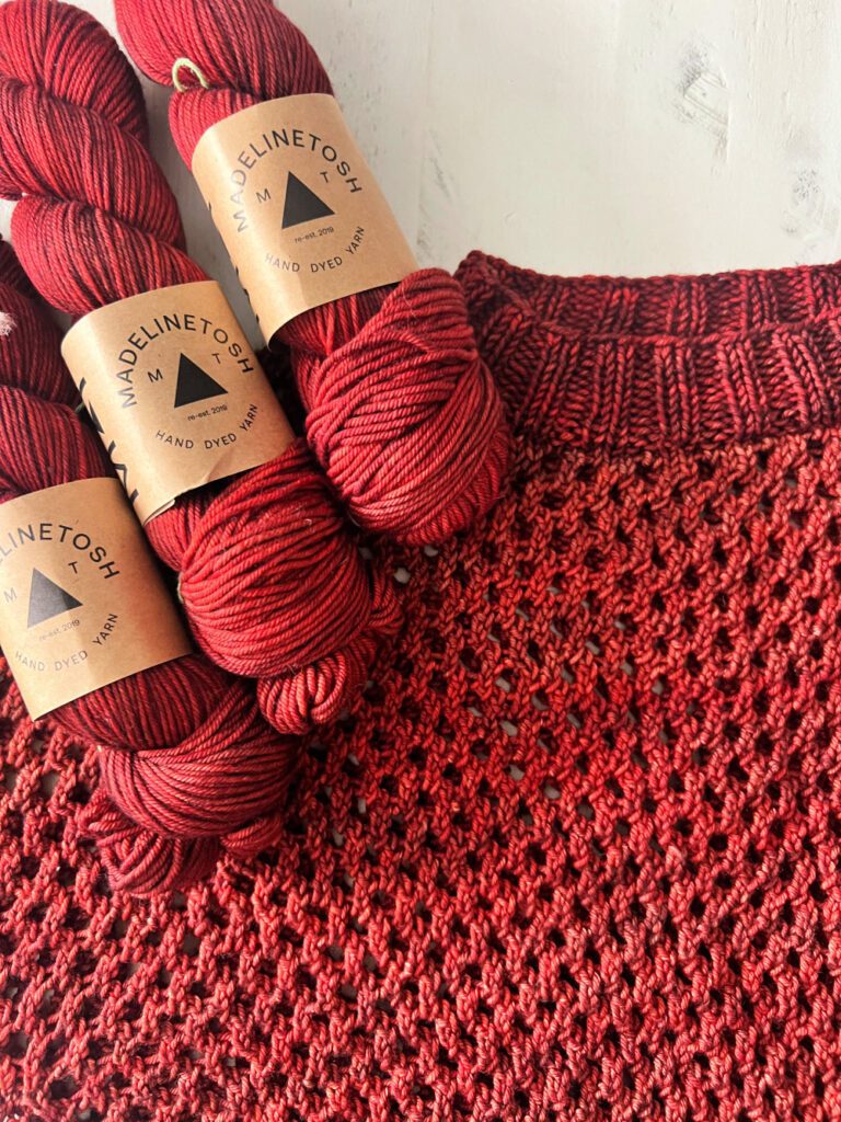 A must-have Chic Sandra creation, this cropped knit sweater is made with deep red yarn and displayed alongside three skeins of matching "MadelineTosh Hand Dyed Yarn." Featuring an intricate, textured stitch pattern, the garment stands out beautifully against a light background. -Marly Bird