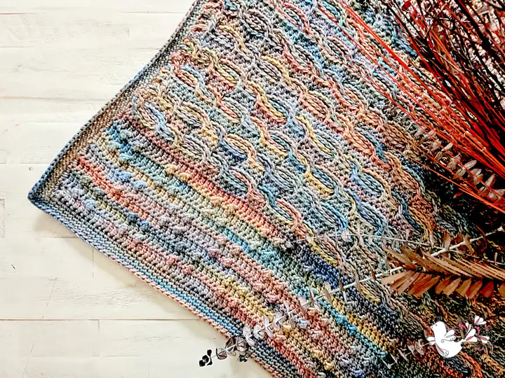 A multicolored crocheted blanket with intricate patterns, inspired by free knitting and crochet patterns, is draped on a light wooden floor. Brown dried foliage lies partially on top of the blanket near one corner. -Marly Bird