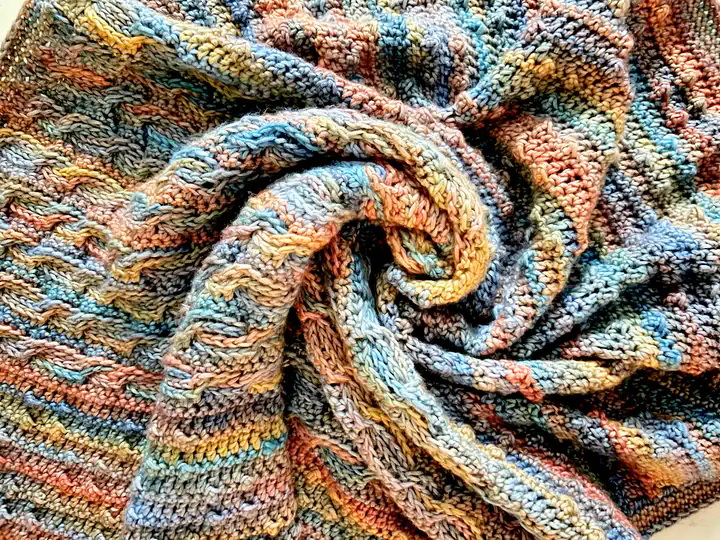 A close-up of a multicolored knitted blanket with textured patterns. The fabric, inspired by free knitting and crochet patterns, is arranged in a loose spiral, showcasing shades of blue, orange, yellow, and beige woven together. -Marly Bird