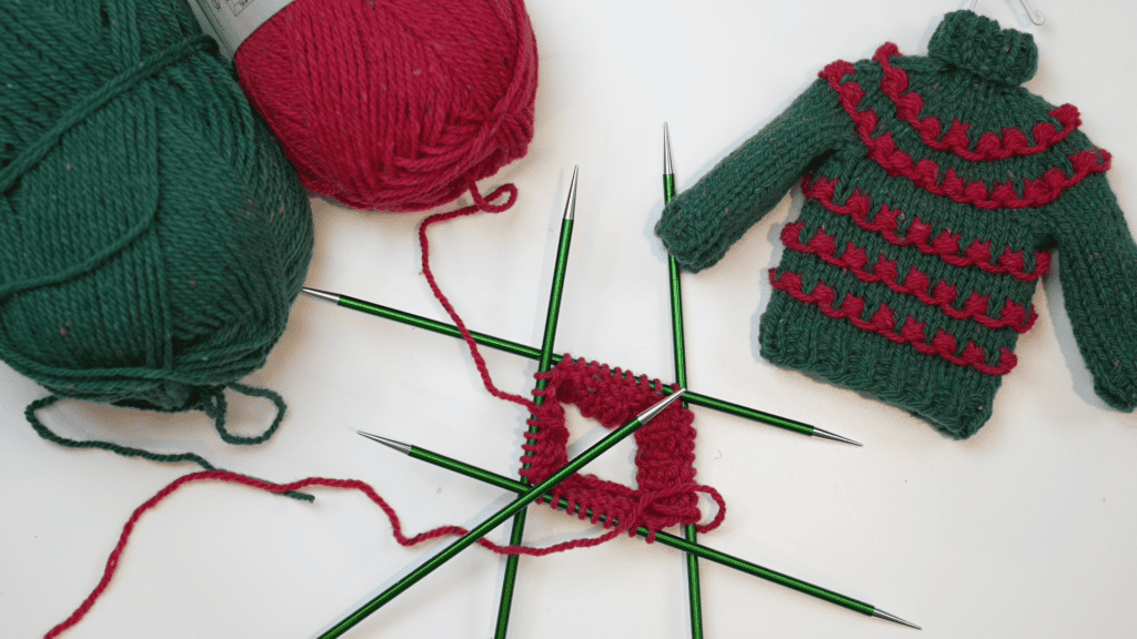 A cozy knitting scene features two balls of yarn, one red and one green. A small Christmas knit sweater ornament with festive red patterns is displayed next to green knitting needles, accompanied by a delightful free pattern for your crafting pleasure. -Marly Bird