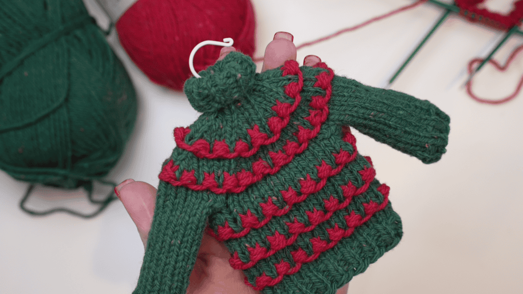 A person's hand holds a small, intricately knitted Christmas sweater in green and red. Balls of yarn and knitting needles are visible in the background, suggesting a festive creation from a free pattern. -Marly Bird