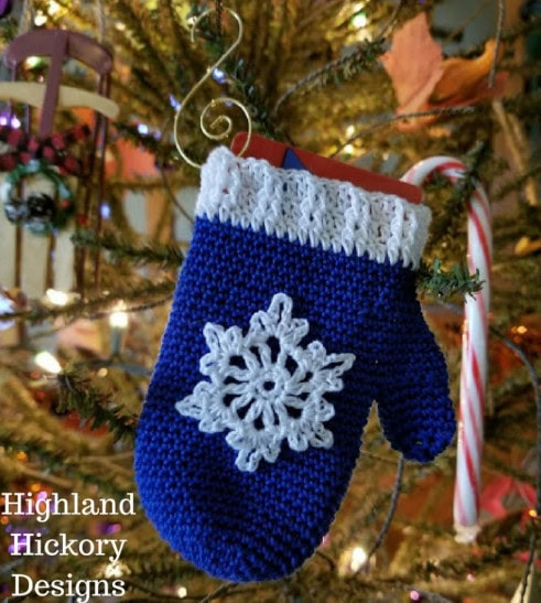 A blue crocheted mitten with a white snowflake decoration hangs on a Christmas tree, serving as festive gift card holders. A candy cane and a card peek out from the mitten. Text reads "Highland Hickory Designs. -Marly Bird