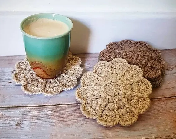 A light green ceramic mug with a latte sits on a beige crocheted coaster. Next to it, two stacks of flower-shaped crocheted coasters in beige and brown—perfect for your 24 days of quickies—rest on a wooden surface. -Marly Bird