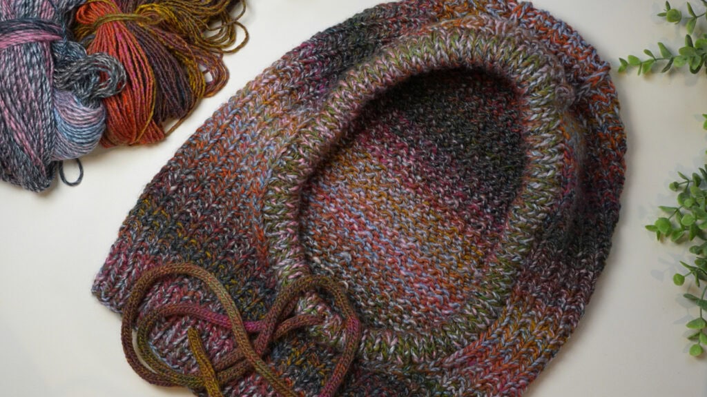 A knit cowl with multicolored stripes and a drawstring sits by matching yarn; leafy plants border the cozy tabletop scene.