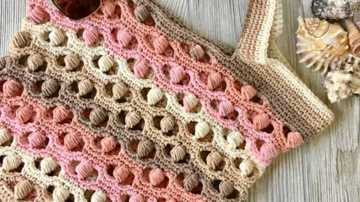 Crochet tote bag with bobble stitch texture in cream, pink, and brown yarn, displayed beside sunglasses and seashells on wood.