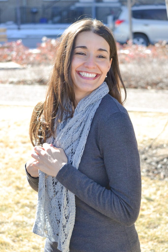 A gray knit lace scarf with a delicate, openwork stitch is modeled outdoors, showing its texture and drape in sunlight.