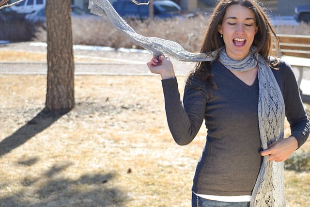 A woman models a knit lace shawl, showing off its airy texture and stitch detail as she drapes it over her shoulder outdoors.
