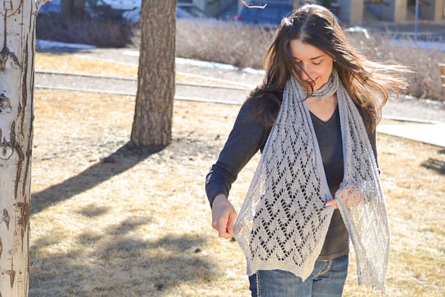 A woman models a light gray lace shawl, showing its delicate stitchwork and airy texture while standing on dry grass.