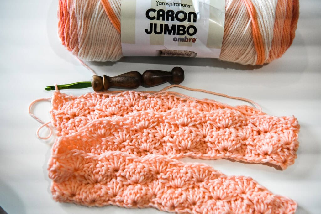 A partially crocheted pink and white scarf using an easy crochet shell stitch pattern, with a green-handled crochet hook and a skein of Caron Jumbo ombre yarn on a white surface. -Marly Bird