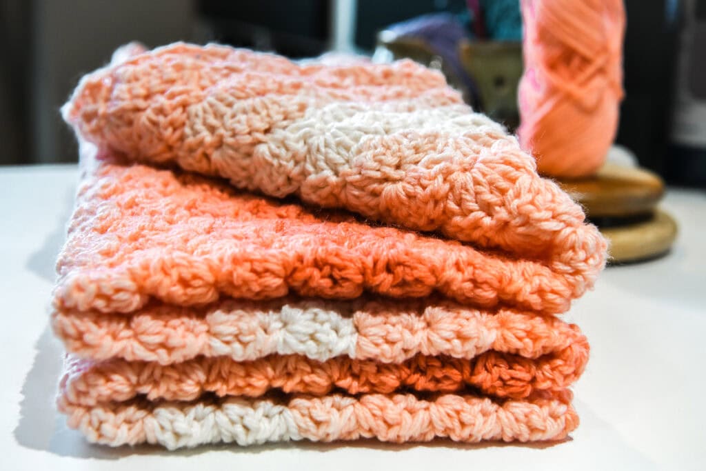 Three neatly folded, crocheted cloths in shades of pink and white—crafted with an easy crochet shell stitch pattern—sit on a white surface, with a ball of yarn and blurred objects in the background. -Marly Bird