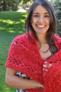 Woman models a red crochet shawl with visible stitch detail over a floral dress, standing on grass with trees behind her.