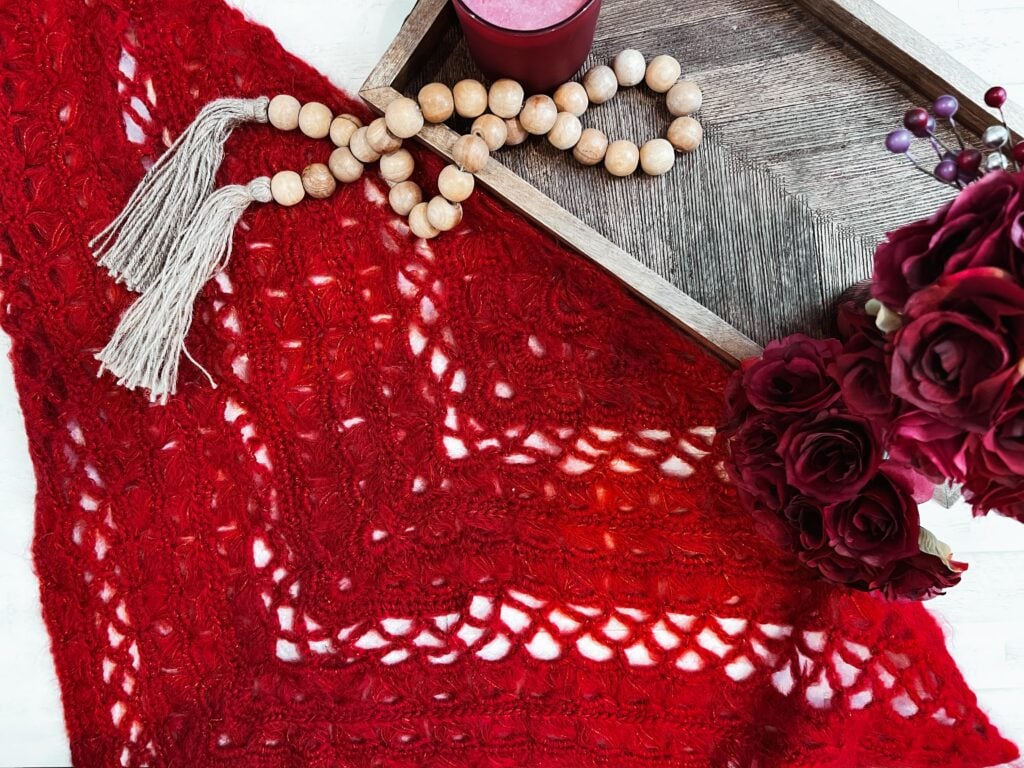 Red broomstick lace crochet shawl showing textured stitches, displayed beside decor on a white background.