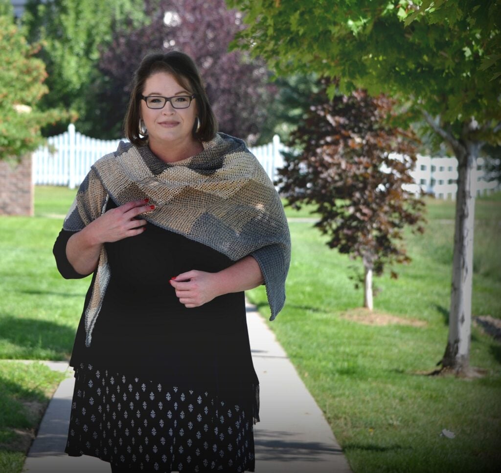 Woman models a crochet wrap shawl over a black dress; shawl shows openwork stitch detail. Trees and fence behind her.