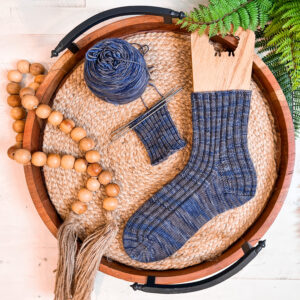 A hand-knitted blue sock and a partially finished sock on needles, with fine stitch detail, displayed on a woven mat in a tray.