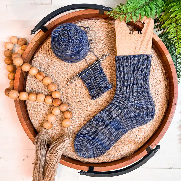 A knitted blue sock on a wooden sock blocker, a matching yarn ball, double-pointed needles with unfinished work, and a bead garland rest on a woven tray by green fern leaves—inviting inspiration for free knitting and crochet patterns. -Marly Bird