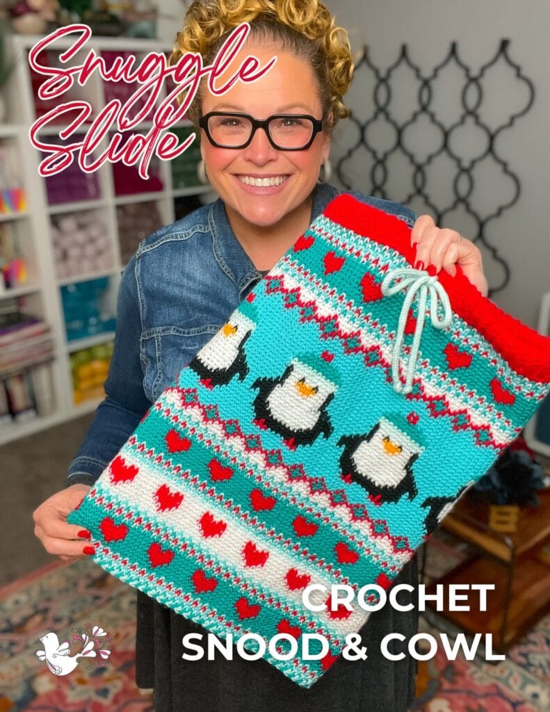 A smiling woman with glasses holds a colorful crocheted snood and cowl featuring penguins and red hearts from Jolly Holiday vol 3. The text says “Snuggle Slide” and “Crochet Snood & Cowl.” Shelves with yarn appear in the background. -Marly Bird