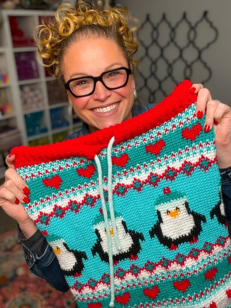 A woman shows a knitted piece with penguin motifs and Split SC colorwork in turquoise, red, white, and black; yarn shelves behind.