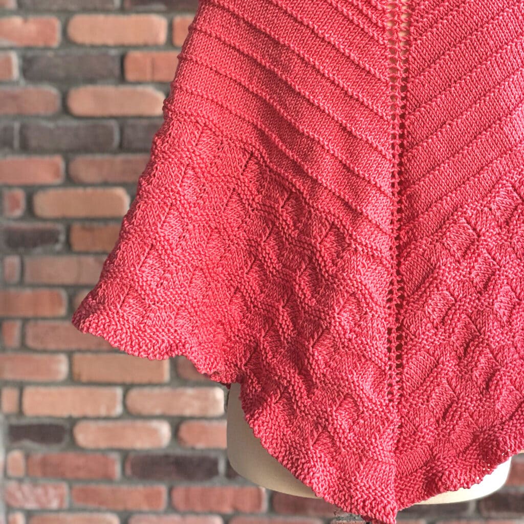 Red knitted shawl featuring textured diamond patterns and diagonal ribbing, shown on a mannequin before a brick wall.