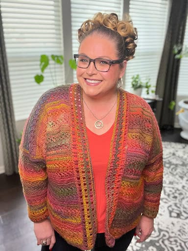 A woman is smiling while wearing glasses and a Sookie Crochet Cardigan over an orange top. Her hair is styled in curls. She stands in a room with large windows and plants, where the patterned rug on the floor complements her vibrant look. -Marly Bird