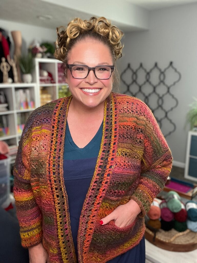 A smiling woman with curly hair and glasses stands indoors, wearing a colorful crocheted cardigan over a blue top. Behind her are shelves with yarn, books, and craft supplies in a cozy, well-lit room. -Marly Bird