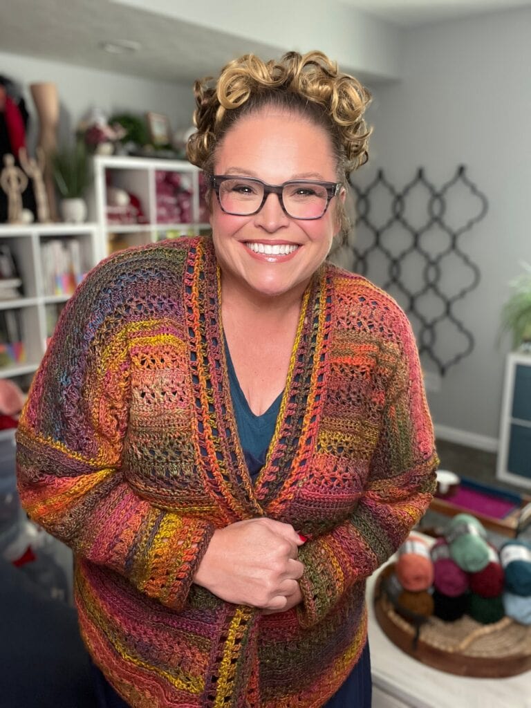 A smiling woman with curly hair and glasses wears a multicolored, crocheted cardigan. She stands in a cozy, well-lit room with yarn, crochet supplies, and bookshelves in the background. -Marly Bird