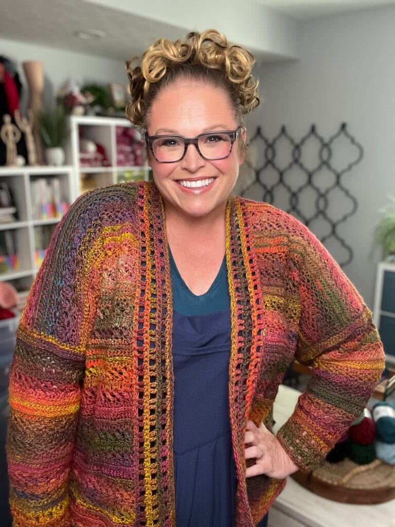 A smiling woman with curly hair and glasses wears a colorful crochet cardigan over a blue top, standing in a cozy craft room with yarn, bookshelves, and plants in the background. -Marly Bird