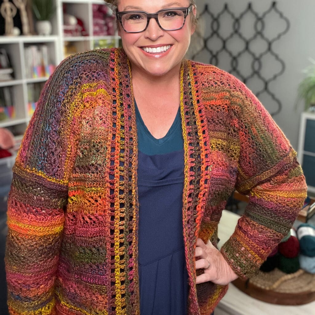 Person wears a vibrant, hand-crocheted Sookie cardigan; visible yarn shelves highlight the colorful stitch texture and detail.