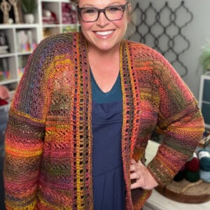 Person wears a vibrant, hand-crocheted Sookie cardigan; visible yarn shelves highlight the colorful stitch texture and detail.