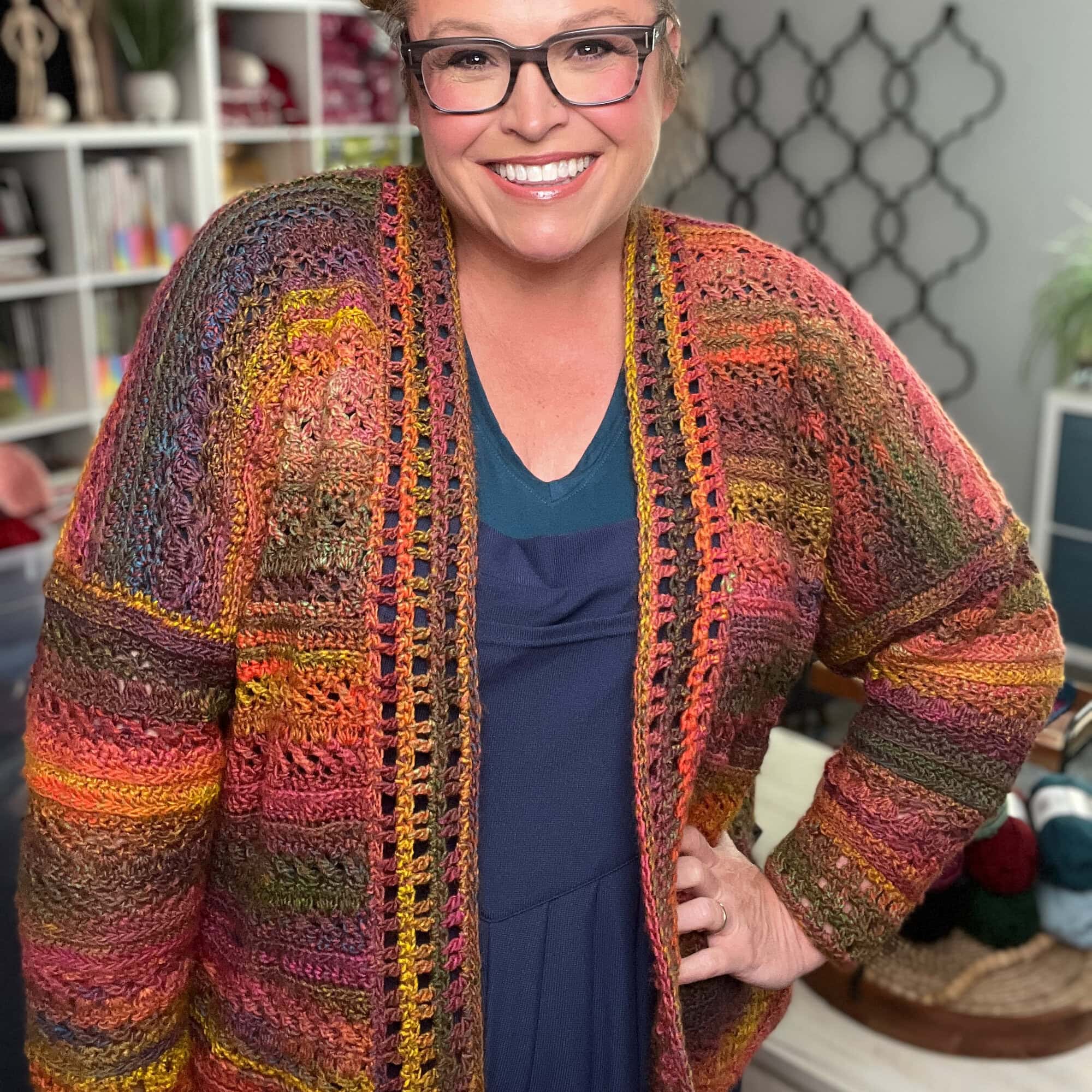 Person wears a vibrant, hand-crocheted Sookie cardigan; visible yarn shelves highlight the colorful stitch texture and detail.